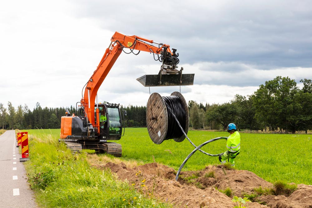 Orange grävmaskin lägger kabel i dike medan arbetare i reflexkläder övervakar arbetet vid landsväg