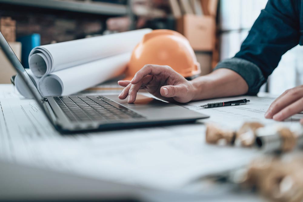 Ingenjör som arbetar vid laptop med ritningar, bygghjälm och tekniska verktyg på skrivbord